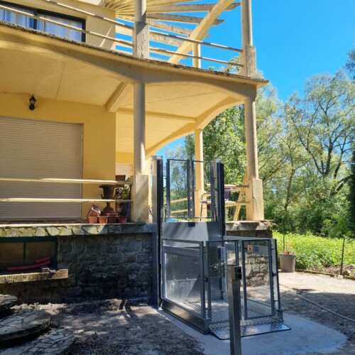 vue de côté de l'élévateur vertical , plateau antidérapant au sol, villa provençale à pergola blanche et escalier courbe en pierre en arrière-plan, ciel bleu