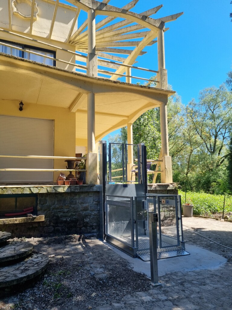 vue de côté de l'élévateur vertical , plateau antidérapant au sol, villa provençale à pergola blanche et escalier courbe en pierre en arrière-plan, ciel bleu