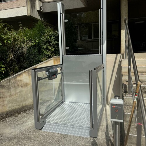 vue de face de l'élévateur vertical aluminium et vitré, plateau antidérapant déployé, escalier en béton brut et bâtiment de bureaux en arrière-plan, borne de commande à droite