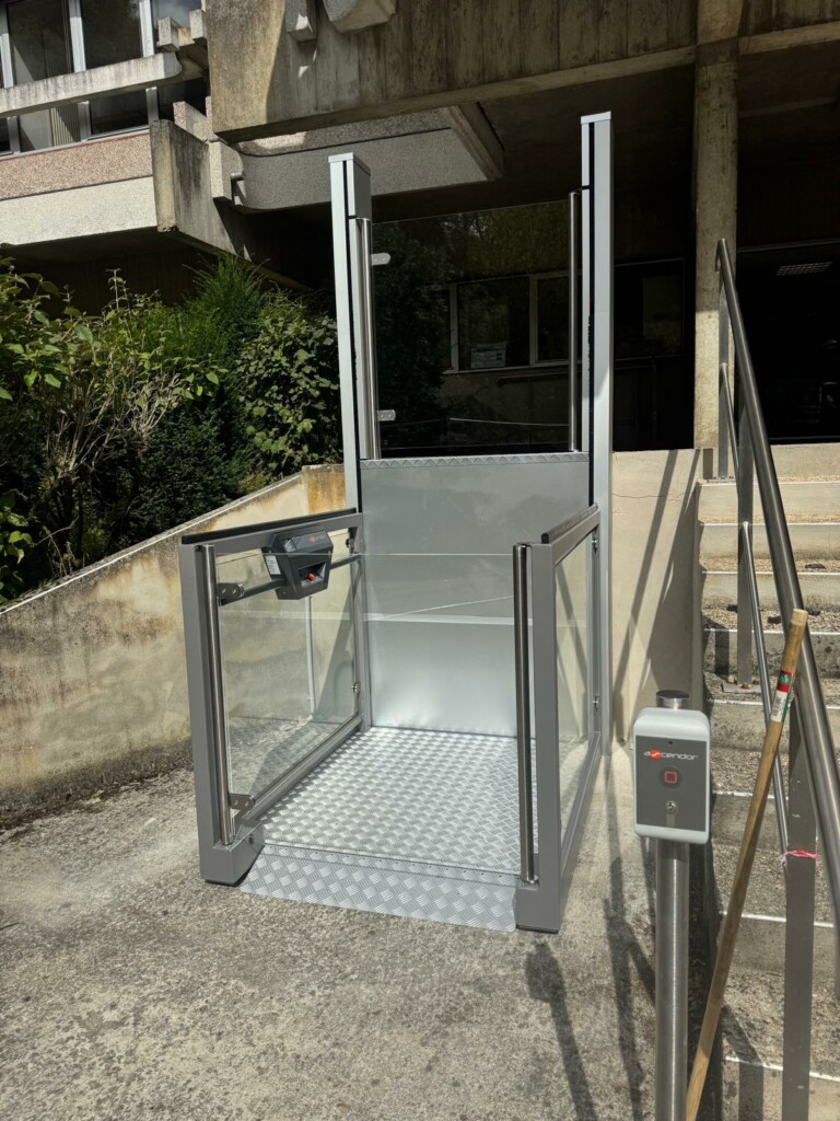 vue de face de l'élévateur vertical aluminium et vitré, plateau antidérapant déployé, escalier en béton brut et bâtiment de bureaux en arrière-plan, borne de commande à droite