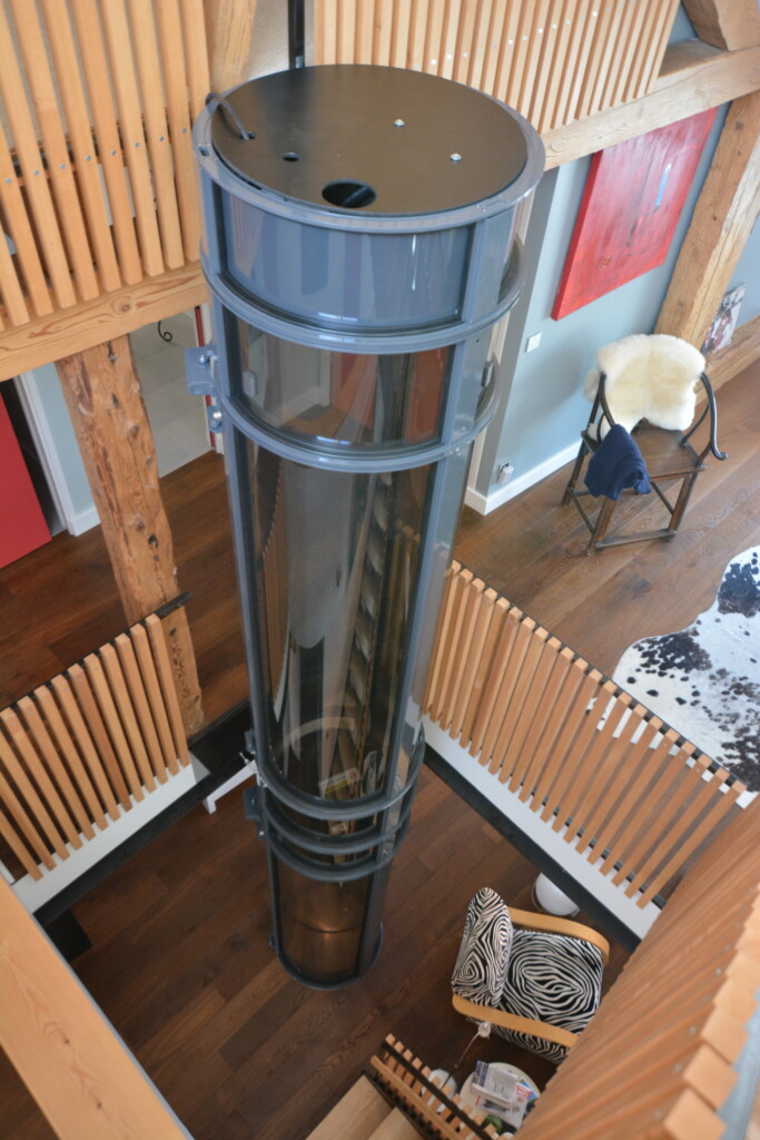 vue plongeante depuis la mezzanine sur l'ascenseur cylindrique transparent, bois naturel blond, parquet, garde-corps et œuvre d'art rouge en arrière-plan