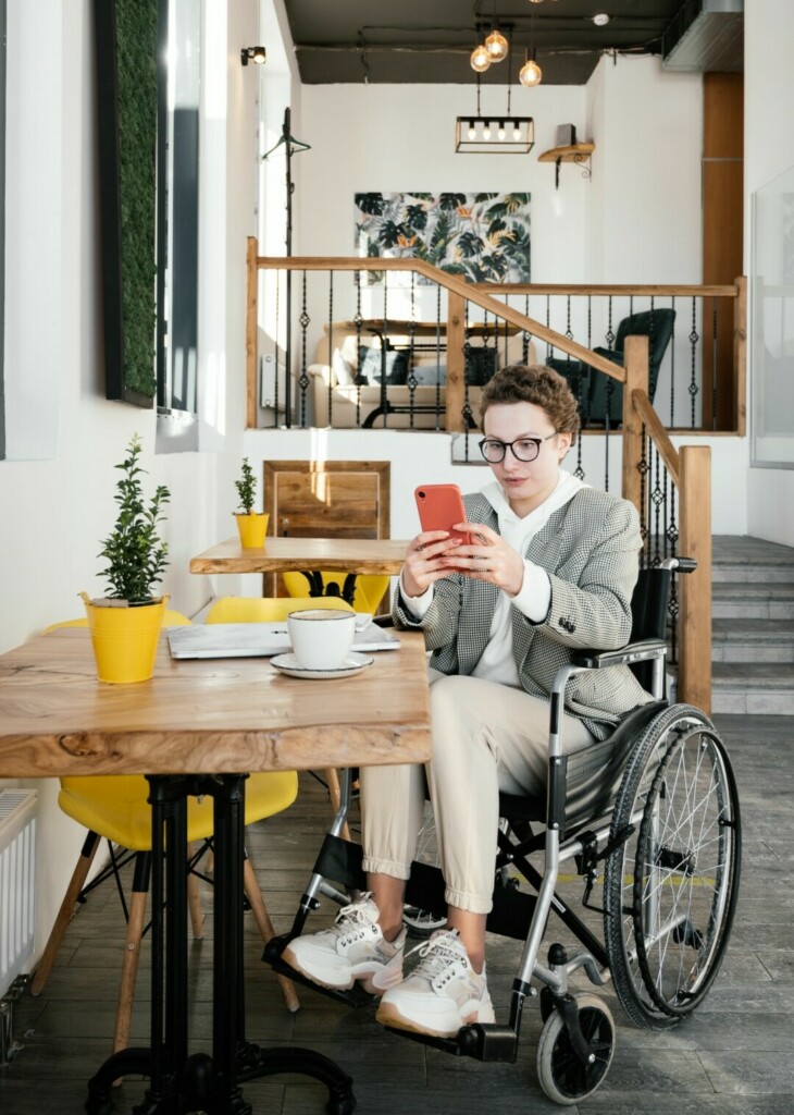 femme installee dans son fauteuil roulant a son domicile sur son telephone avec en fond d'image quelques marches pour acceder au salon