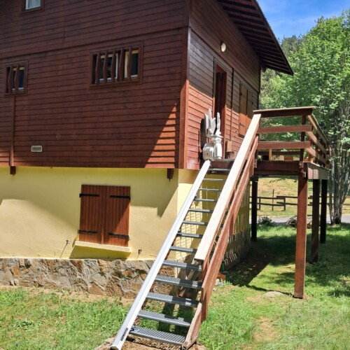 vue d'ensemble du chalet en bois brun, escalier extérieur en caillebotis métallique, rail aluminium sur toute la longueur, fauteuil replié en haut, prairie verte et ciel bleu en arrière-plan