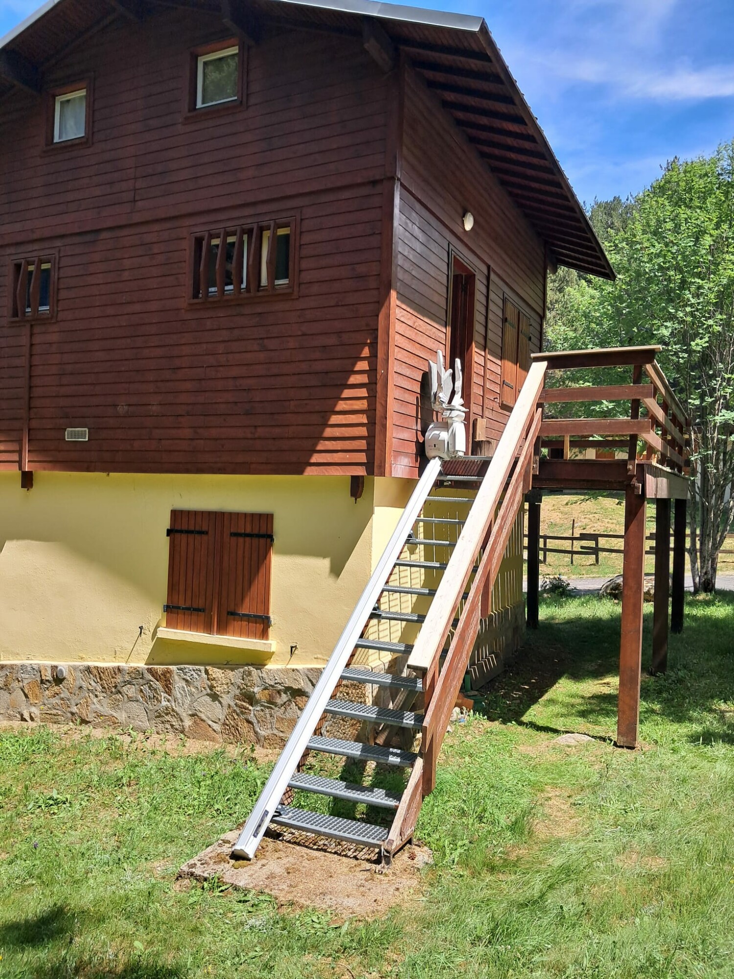 vue d'ensemble du chalet en bois brun, escalier extérieur en caillebotis métallique, rail aluminium sur toute la longueur, fauteuil replié en haut, prairie verte et ciel bleu en arrière-plan