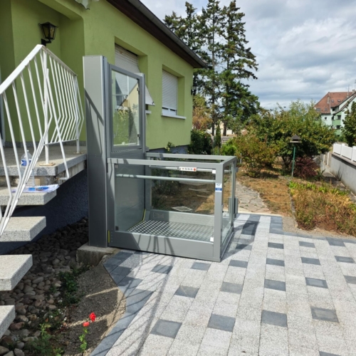 photo principale — vue d'ensemble depuis le jardin : façade verte, terrasse en pavés bicolores neufs, élévateur Sigma gris anthracite avec parois vitrées, escalier en granit à gauche, jardin fleuri